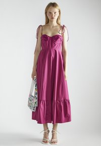 Magenta katoenen jurk met een aangesloten lijfje, volantrand en verstellbare schouderbanden. Model houdt een bewerkte handtas met bloemen en draagt witte hakken.