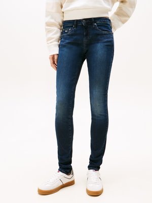 Persona che indossa jeans slim-fit blu scuro, scarpe da ginnastica bianche con suole in gomma e una maglietta a maniche lunghe di colore chiaro, in piedi davanti a uno sfondo bianco.