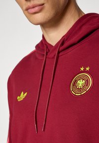 Maroon hoodie featuring a yellow embroidered German football logo and an Adidas logo in yellow, with drawstrings and a textured fabric.