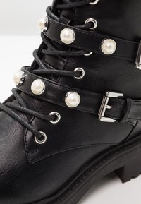 Stivale alla caviglia in pelle nera con lacci, fibbie argentate e cinturini decorativi con perle bianche rotonde.
