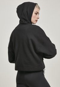 Felpa nera con cappuccio, caratterizzata da un design accorciato, una texture morbida e un dettaglio con lacci sul bordo. Le maniche sono ampie e rilassate.