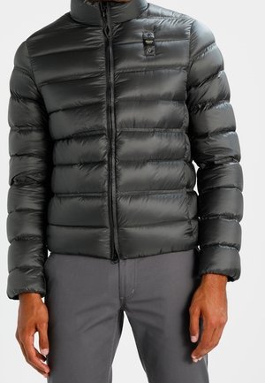 Černá prošívaná péřová bunda s předním zipem, kterou nosí osoba oblečená v šedých kalhotách, ruce volně podél těla.