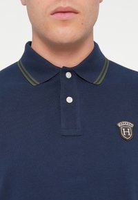 Sötétkék pólóing texturált anyagból, két gombos nyakrésszel, zöld részletekkel a galléron és hímzett logóval a mellkason.