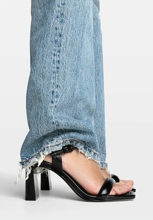 Black heeled sandals with a wide strap and adjustable buckle. Paired with light blue, distressed denim jeans featuring frayed hems.