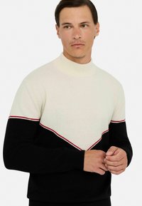 Strickpullover mit hohem Kragen, cremefarbener Oberseite, schwarzen Ärmeln und einem roten Akzentstreifen, der in V-Form über die Brust verläuft.