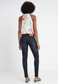Top halter blanco con estampado floral colorido, combinado con jeans ajustados oscuros. El modelo lleva zapatos de tacón abiertos verdes. La textura de la tela parece suave.