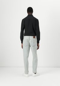 Camisa negra de manga larga con cuello puntiagudo, combinada con jeans de mezclilla azul claro y zapatillas blancas, vista desde la parte trasera.