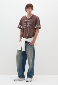 Camiseta de punto marrón con texto blanco y mangas a rayas, combinada con jeans de mezclilla azul claro, un suéter blanco atado a la cintura y zapatillas grises.