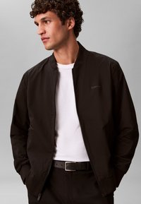 Veste bomber noire avec fermeture éclair à l'avant, poignets et col côtelés. Portée sur une chemise blanche, avec un logo discret sur le côté gauche de la poitrine.