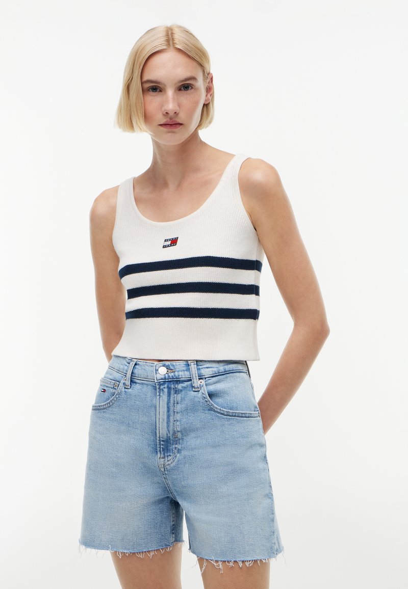 Γυναίκα που φοράει λευκή αμάνικη κορυφή crop top με ναυτικά μπλε ρίγες και ανοιχτό μπλε τζιν σορτς ψηλόμεσο με φθαρμένες άκρες, στέκεται μπροστά σε λευκό φόντο.