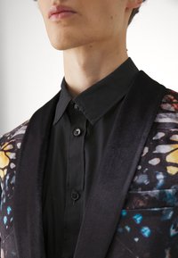 Tailored jacket with a black velvet lapel, featuring a colorful butterfly print. Black button-up shirt underneath, smooth texture.