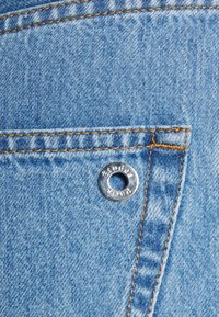 Světlá modrá džínová látka s stříbrným průchodem a kontrastním hnědým prošitím podél švu, která ukazuje texturovaný povrch.