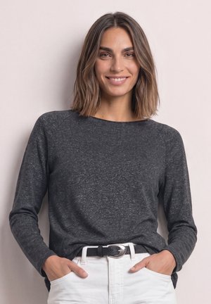 Femme aux cheveux bruns mi-longs souriante, portant un haut à manches longues gris foncé et un pantalon blanc avec une ceinture noire, debout contre un mur clair.