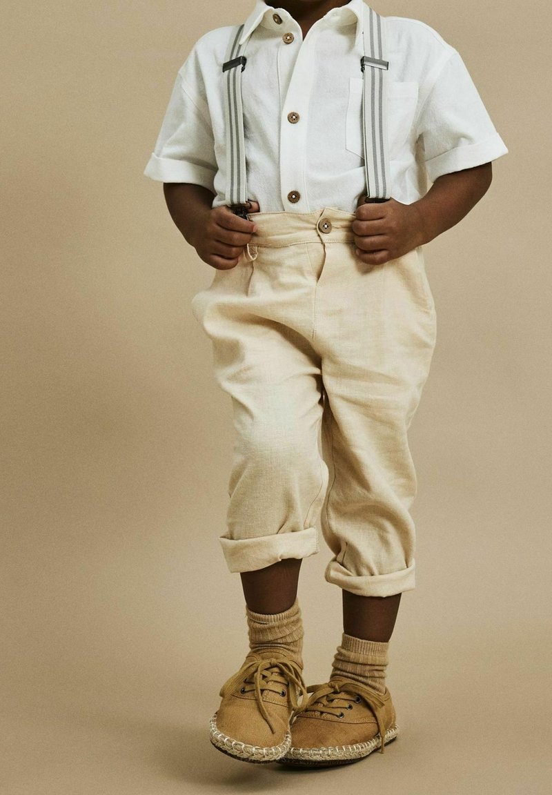 Jasno beżowe spodnie z podwiniętymi nogawkami, biała koszula z krótkim rękawem z drewnianymi guzikami, szare szelki oraz brązowe buty z plecioną podeszwą.