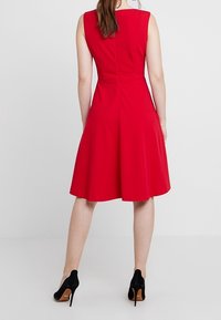 Rode mouwloze jurk met een uitlopende rok. Het materiaal lijkt glad en zit aangesloten op de taille. Gecombineerd met zwarte hoge hakken.