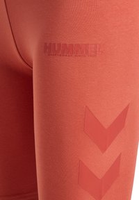 Gros plan sur des leggings rouges avec le logo "HUMMEL" embossé et deux motifs chevrons vers le bas sur la cuisse.