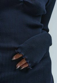 Granatowy, prążkowany sweter z długimi rękawami, z postrzępionymi mankietami. Materiał ma miękką teksturę i dopasowany krój, podkreślający kontury ciała.