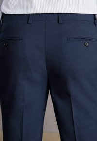 Marineblaue Hose aus strukturiertem Stoff, mit zwei Gesäßtaschen mit schwarzen Knöpfen und tailliertem Schnitt. Gerades Bein.