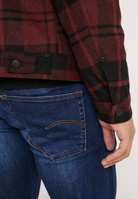 Rød og sort ternet flaneljakke med sorte knapper, kombineret med blå denimjeans med orange syninger og et karakteristisk lomme-design.