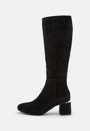 Schwarze Wildlederstiefel in Knielänge mit runder Zehe, klobigem Absatz und metallischem Akzent am Fußbereich. Glatte Textur und figurbetonte Form.
