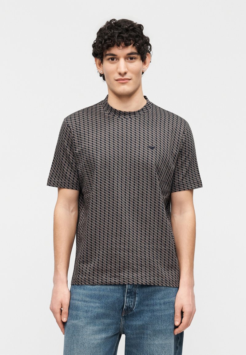 Νεαρός άντρας με σγουρά μαλλιά που φοράει κοντομάνικο πουκάμισο με μαύρο και μπεζ σχέδιο και μπλε τζιν, στέκεται μπροστά σε απλό λευκό φόντο.