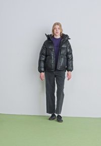 Čierna puffa bunda s lesklým povrchom, kapucňou a prednými vreckami, nosená cez fialovú košeľu a spárovaná s čiernymi nohavicami a topánkami.