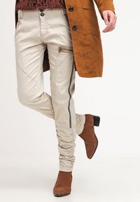Beżowe spodnie w slim fit z zamkiem błyskawicznym, czarnymi paskami po bokach i marszczonym detalem przy kostce, w połączeniu z brązowymi botkami na obcasie.