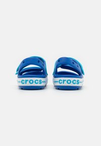 Zuecos azules de Crocs fabricados con un material de goma texturizada, con una correa trasera y suela blanca. El nombre de la marca "crocs" está impreso en azul en el costado.