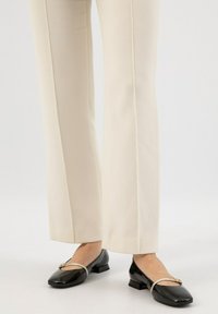 Pantaloni su misura color crema abbinati a mocassini neri in vernice con un accento in oro e design a punta quadrata.