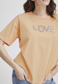 Persikins värinen puuvillapaita, jossa on lyhyet hihat. Paidassa on kirjailtu teksti "LOVE" sinisin ja ruskean värisin kirjaimin. Rentomallinen, jossa on pyöreä kaula-aukko.