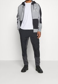 Man in een grijze en zwarte Nike Air rits-jas, een wit T-shirt, zwarte en grijze broeken en zwarte sneakers, staande tegen een witte muur.