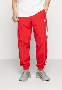 Röda träningsbyxor i lätt material, med vita sidsnor och elastiska mudd vid anklarna. Adidas-logotyp på framsidan.