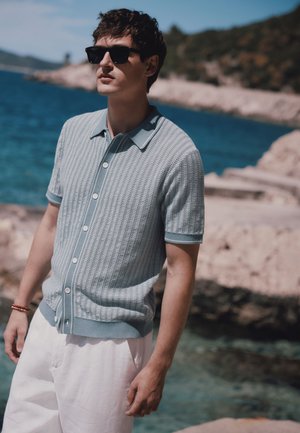 Man wearing sunglasses, light blue short-sleeve knit shirt, and white pants standing by rocky shore with blue sea and hills in background.