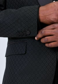 Mustkast mustriga bleiser, millel on sile krae ja kolm musta nööpi mansetil ning üks tasku ees.