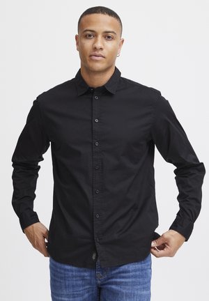 Camisa negra de manga larga y botón, hecha de algodón, con un cuello clásico y placket frontal con botones, llevada sobre unos jeans azules.