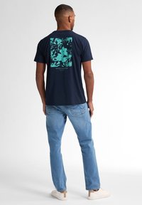 T-shirt bleu marine avec un graphique tropical en vert menthe et du texte au dos, associé à un jean bleu clair et des baskets blanches.