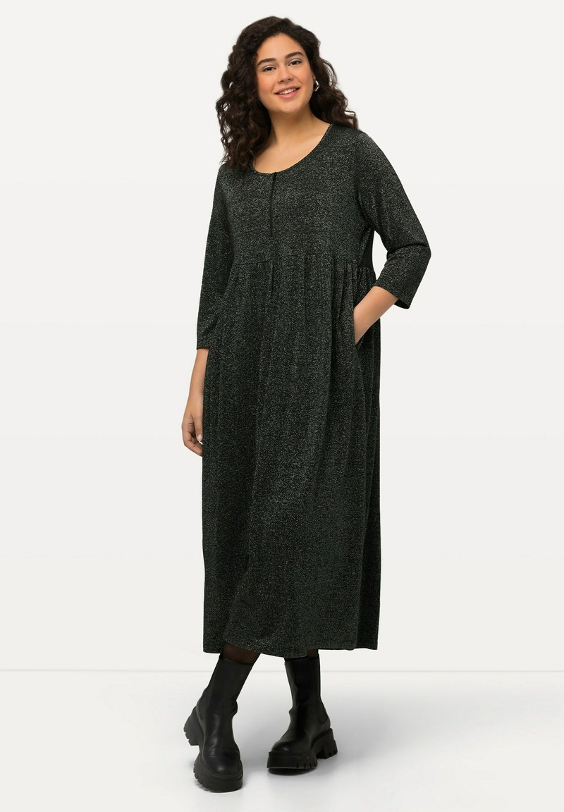 Robe noire scintillante avec un décolleté arrondi, des manches trois-quarts et des poches latérales. Design long et fluide, associée à des bottes noires épaisses.
