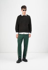 Maglione nero a costine sopra una camicia bianca con colletto, abbinato a pantaloni da jogging verdi con strisce bianche e scarpe chunky nere.