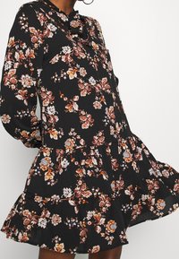 Czarna sukienka w kwiaty z długimi rękawami, warstwami falbanek i paskiem w talii. Posiada pomarańczowe i białe wzory kwiatowe na teksturowanej tkaninie.