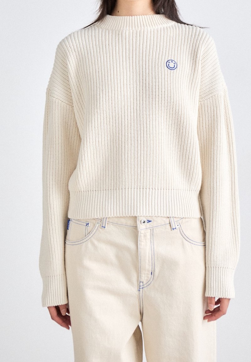 Pull en maille crème avec une texture côtelée, épaules tombantes et un petit emblème bleu de visage souriant sur le côté gauche. Longueur raccourcie.