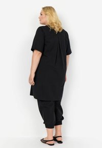 Tunika nera con maniche corte, dettaglio plissettato sulla parte posteriore, abbinata a pantaloni neri con polsini elasticizzati e sandali aperti davanti. Tessuto dalla texture liscia.