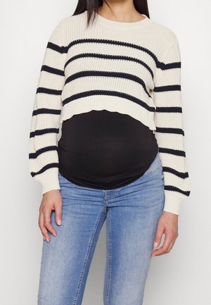 Femme enceinte portant un pull court blanc à rayures noires, un haut de maternité noir et un jean bleu clair, debout devant un fond uni.