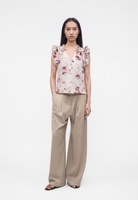 Femme debout portant un chemisier sans manches à volants à motif floral, un pantalon beige à jambes larges et des sandales noires à bout ouvert sur fond uni.