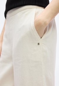 Χέρι στην τσέπη ανοιχτό μπεζ παντελονιού με μικρό λογότυπο DKNY στην τσέπη, σε συνδυασμό με μαύρο ριμπ τοπ.