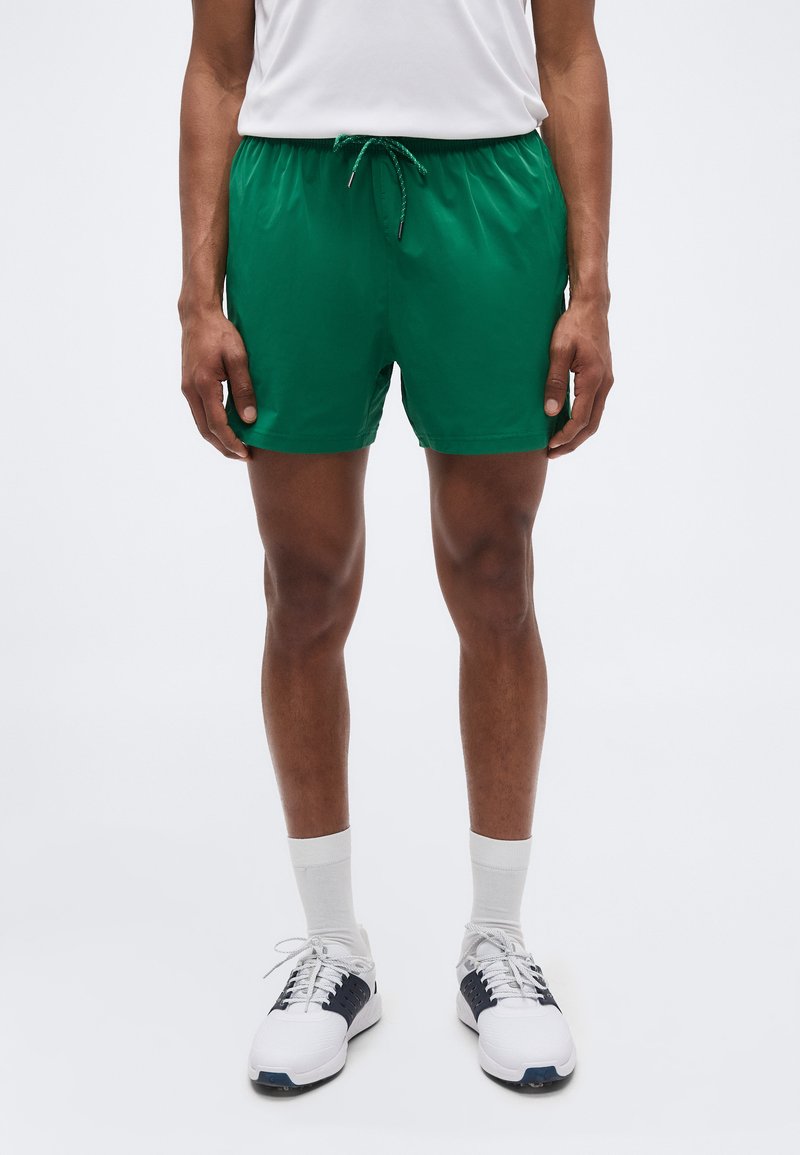 Άνδρας που φοράει πράσινα αθλητικά σορτς, λευκές κάλτσες, λευκά και μαύρα παπούτσια, στέκεται μπροστά σε απλό ανοιχτό φόντο.