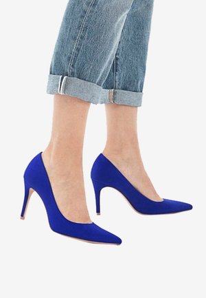 Pieds portant des escarpins pointus bleu vif associés à un jean en denim bleu clair retroussé sur un fond blanc.