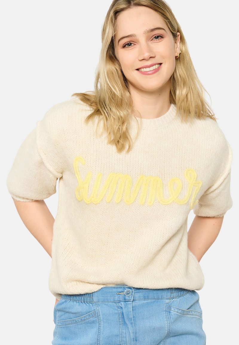 Maglione a maglia color crema con maniche corte arrotolate. Caratterizzato da un design di testo giallo "Estate". Abbinato a pantaloni corti in denim blu chiaro.