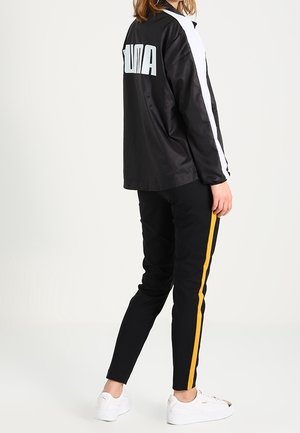 Giacca leggera nera con lati bianchi, con il logo "PUMA" sul retro; abbinata a pantaloni neri con striscia gialla laterale. Sneakers bianche.