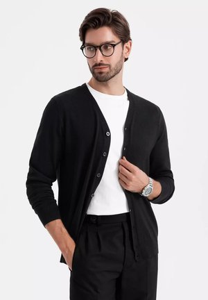 Cárdigan negro hecho de un tejido suave, con un cierre de botones en la parte delantera y puños acanalados. Lleva puesto sobre una camiseta blanca, combinado con pantalones negros.