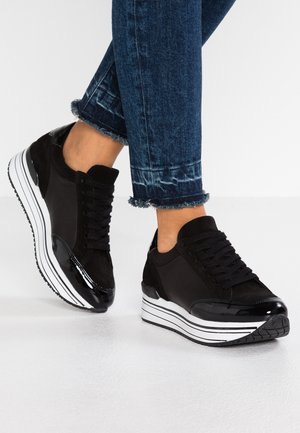 Czarne sneakersy na platformie z białymi paskowanymi podeszwami noszone z podwiniętymi ciemnoniebieskimi dżinsami na białym tle.
